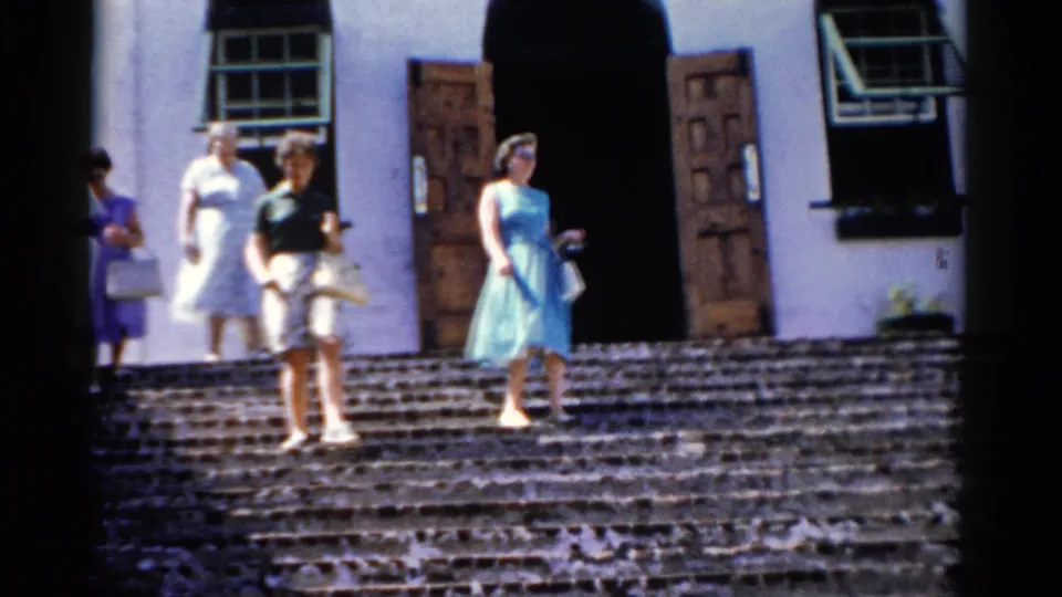 1975 Florida home-movie frame of women in blue, white, and dark tops descending sunlit stone steps outside a white building.