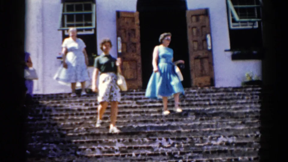 1975 Florida 8mm frame with women in dresses and handbags walking down bright stone steps from a white exterior building.