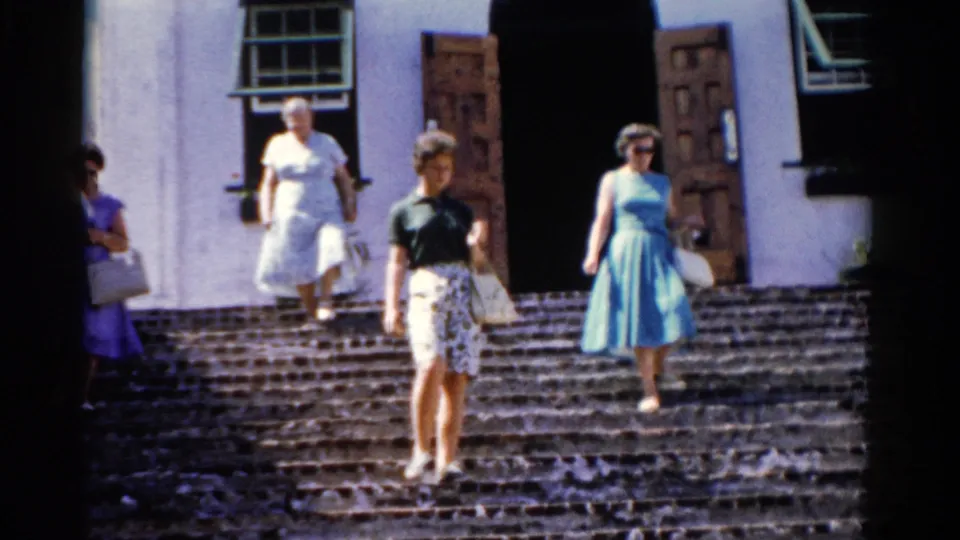 1975 Florida home-movie frame showing women on sunlit stone steps before a white building with open wooden doors.