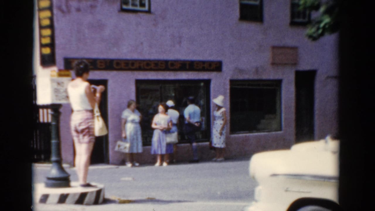 1975 Florida 8mm frame showing a woman with a bag on a striped divider, with people outside The St. Georges Gift Shop behind her.