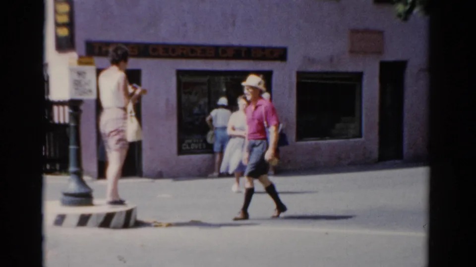 1975 Florida home-movie frame showing a man in a pink shirt crossing in front of a purple gift shop toward a woman on a traffic divider.