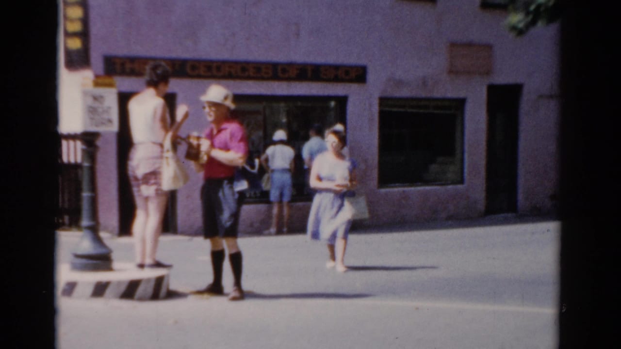 1975 Florida home-movie frame showing pedestrians outside The St Georges Gift Shop, with a woman in pink speaking near a lamppost.