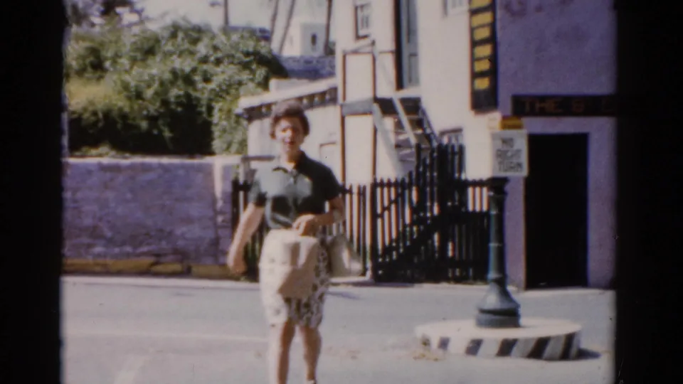 1975 Florida frame of a woman approaching the camera beside The St Georges Gift Shop, a lamppost, fence, and sunlit street.