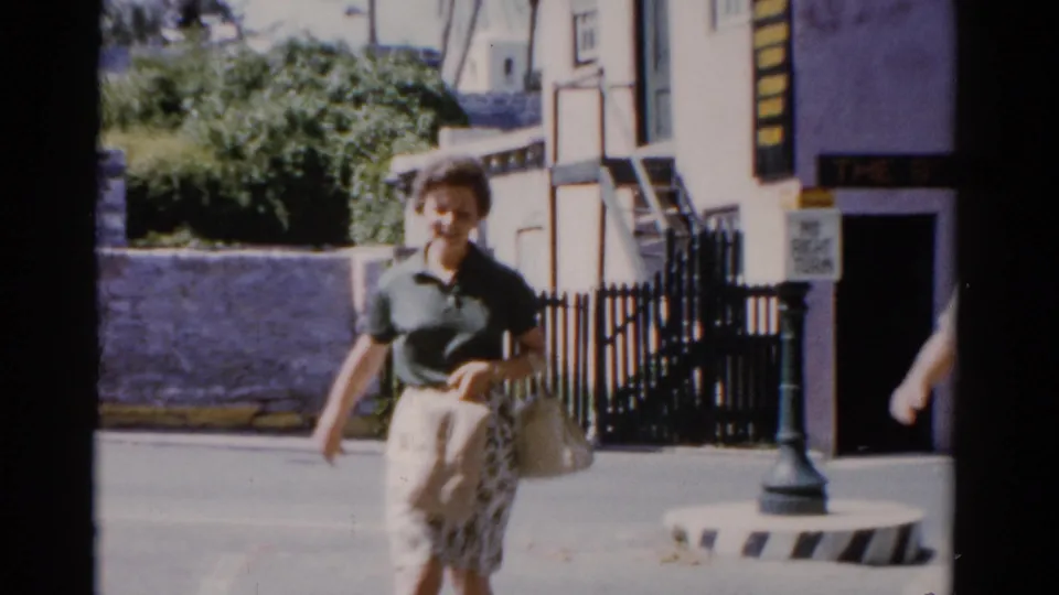 In 1975 Florida home-movie footage, a woman walks outside a pastel building with a black fence and palm-lined background.