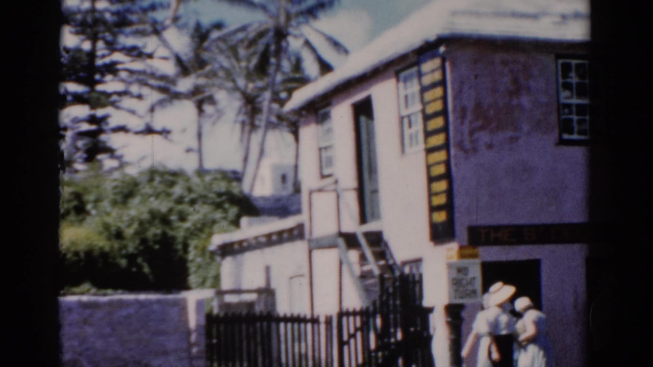 A pastel coastal building appears beside palm trees and a black fence, with two people visible near the entrance area.