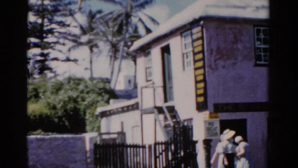 A pastel coastal building appears beside palm trees and a black fence, with two people visible near the entrance area.