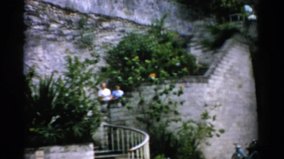 A soft 8mm view shows two adults near the base of a stone stairway with white railing, masonry walls and lush planting.
