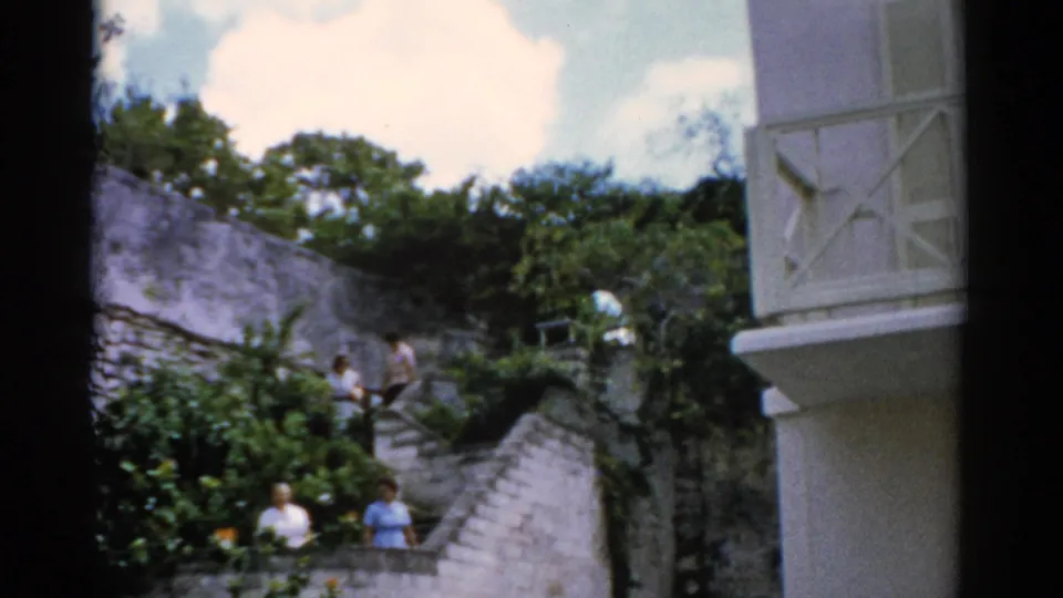 1975 Florida home-movie frame of several adults on stone steps beside lush greenery and a white stucco architectural edge.