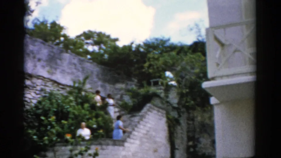1975 Florida home-movie frame showing adults on stone garden steps with tropical planting, stucco walls and a white balcony at right.