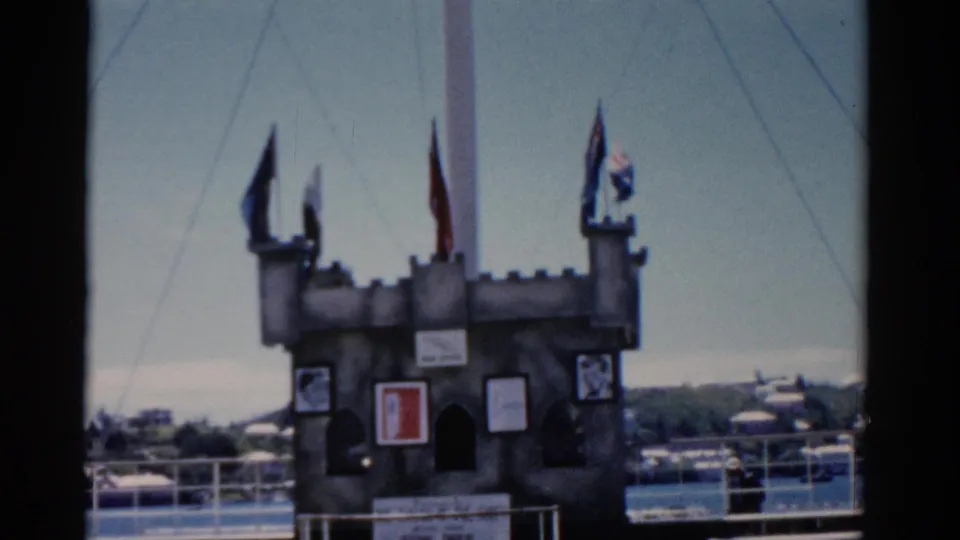 1975 Florida 8mm frame showing a small castle-like dockside attraction with flags, signs, and water beyond.