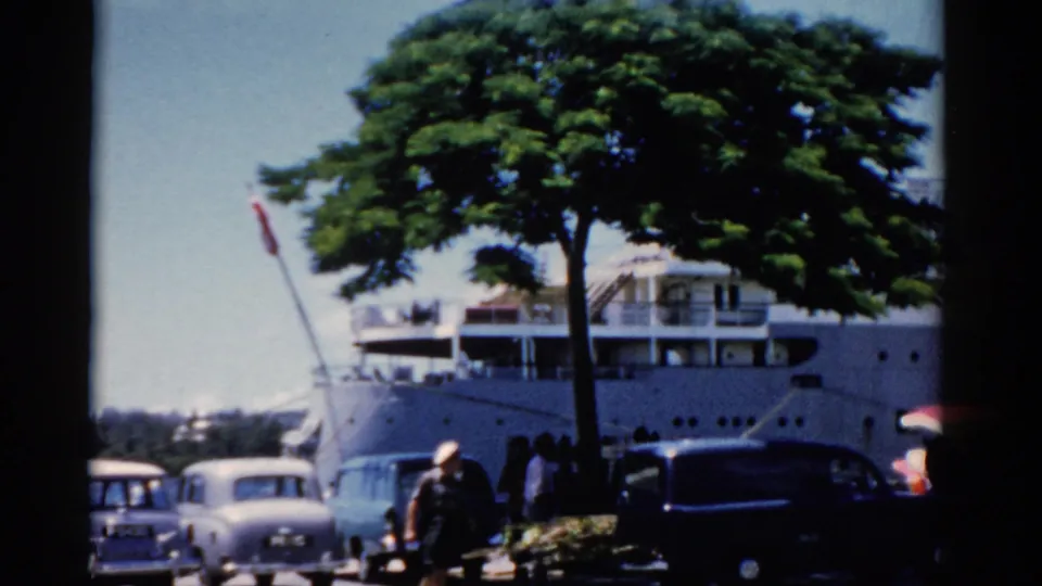 1975 Florida home-movie frame of a docked ship seen from the street with cars, pedestrians, and a large green tree.
