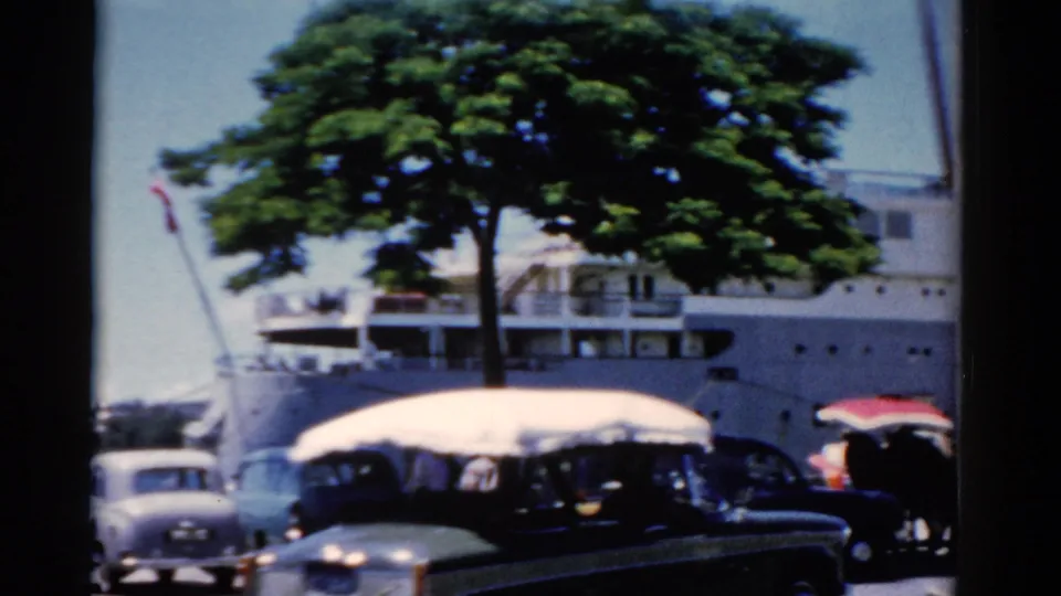 1975 Florida waterfront frame with a dark car, umbrellas, pedestrians, a leafy tree, and a docked white ship behind them.