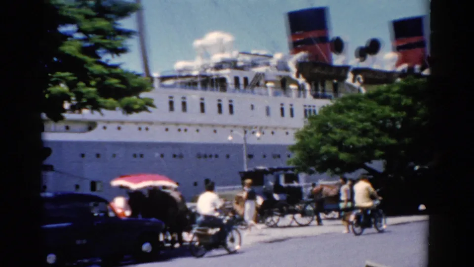 1975 Florida waterfront street scene with a docked passenger ship, horse-drawn carriages, motorbikes, pedestrians, and trees.