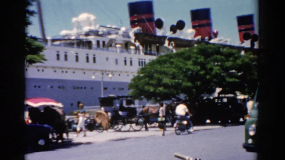 1975 Florida port view with a white passenger ship behind carriages, motorcyclists, cars, trees, and people moving along the street.