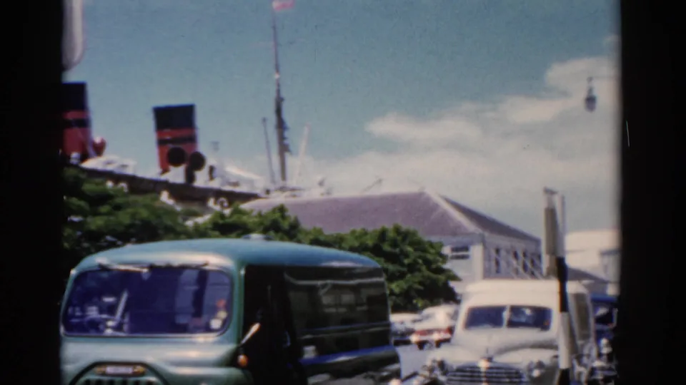 1975 Florida harbor street with a green van, white sedan, pedestrians, lamps, and a large cruise ship visible beyond the roadway.