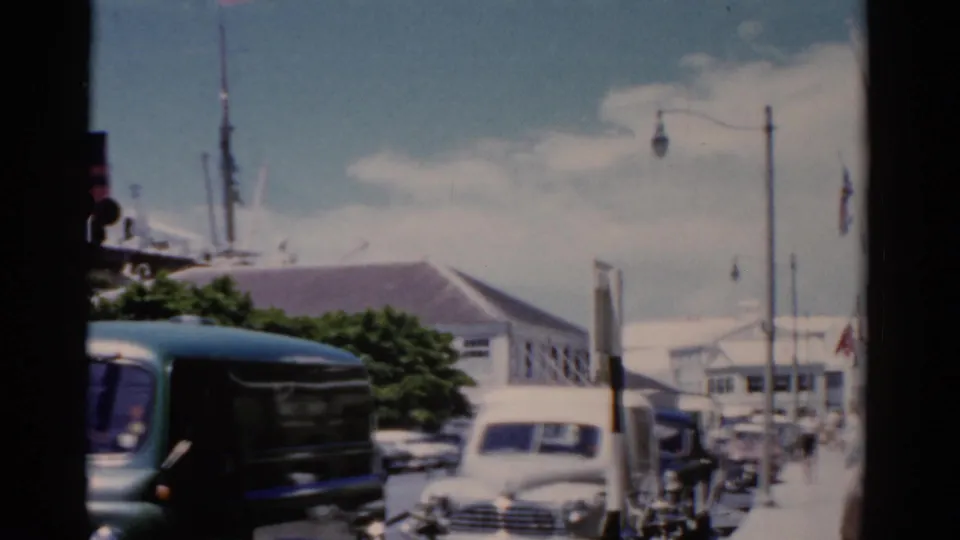1975 Florida port street view with the green van partly leaving frame, a white sedan, pedestrians, lamps, trees, and low waterfront buildings.