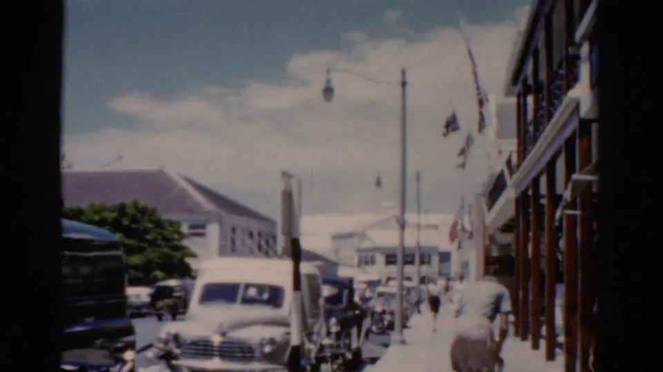 1975 Florida street scene with a white sedan, pedestrians on the sidewalk, flags on a building, street lamps, and harbor buildings in the distance.