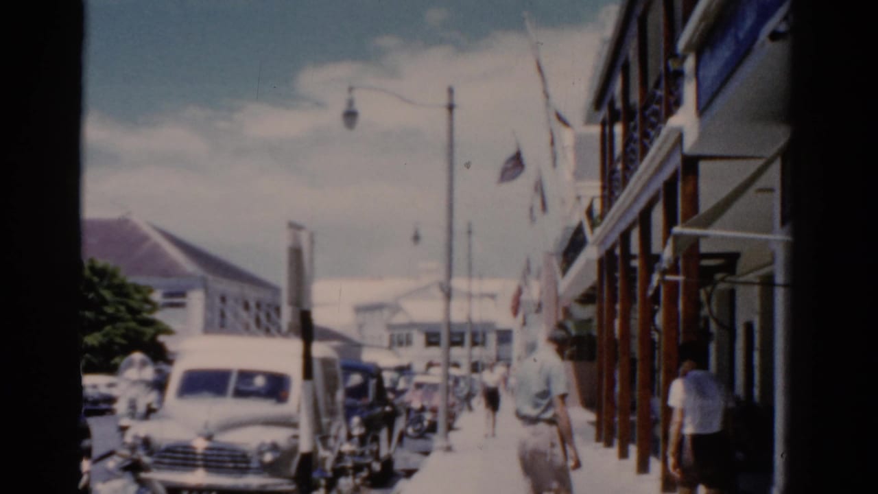 1975 Florida street view with classic cars, pedestrians, flags, and colonial-style buildings seen from handheld 8mm footage.
