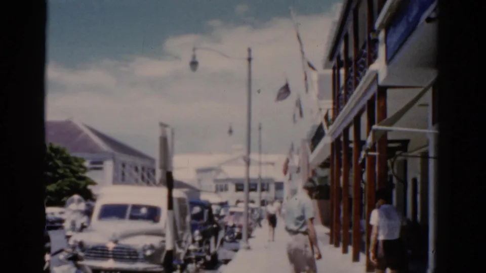 1975 Florida street view with classic cars, pedestrians, flags, and colonial-style buildings seen from handheld 8mm footage.