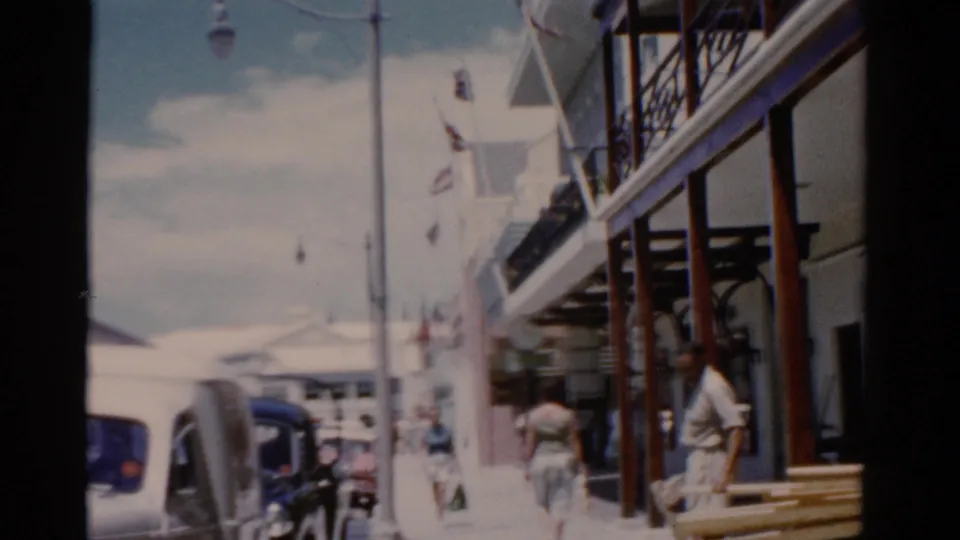 Handheld 8mm view of a Florida sidewalk in 1975 with pedestrians, parked cars, flags, and colonial-style building facades.