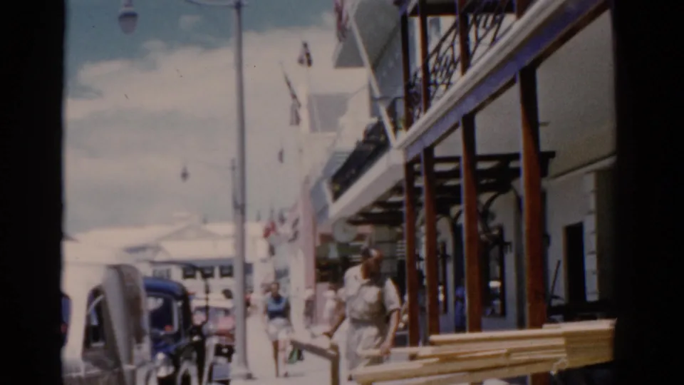 1975 Florida sidewalk frame showing a man carrying wooden planks near parked cars, pedestrians, flags, and raised balconies.