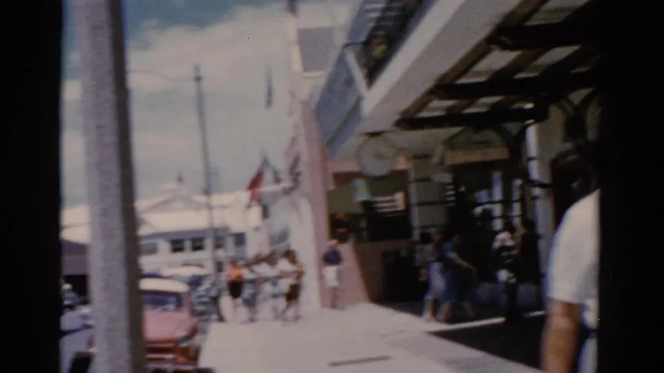 Florida street scene from 1975 with a red car, pedestrians, flags, balcony structures, and strong sun on the sidewalk.