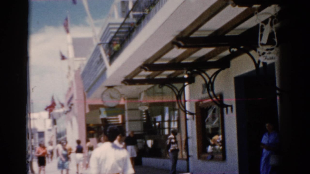 Pedestrians walk along a sunny shop-lined sidewalk with awnings and a hanging clock.