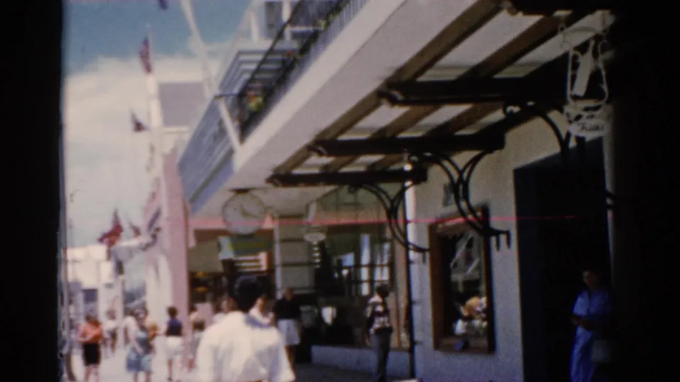 1975 Florida 8mm frame of people walking past storefronts, metal awnings, flags, and a visible hanging clock.