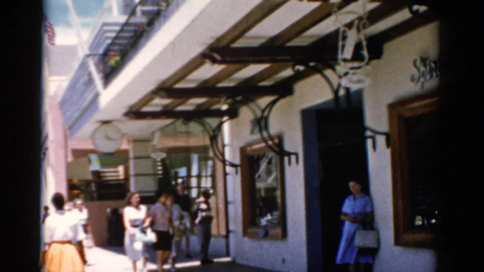 1975 Florida home-movie frame of pedestrians on a shop walkway with awnings, window displays, and a woman standing by the entrance.