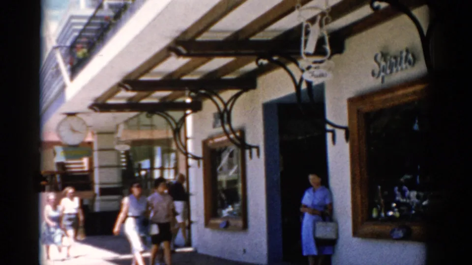 1975 Florida 8mm frame with pedestrians near a shop entrance, visible signage, a hanging clock, and strong midday sunlight.