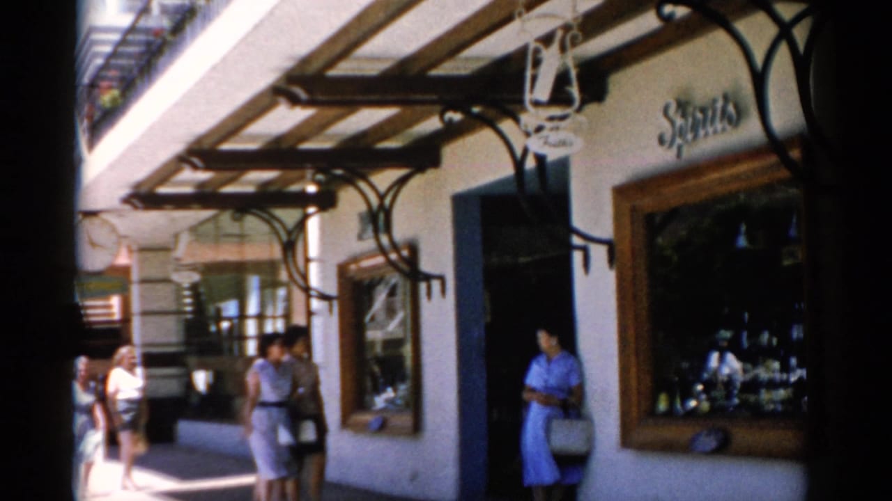 1975 Florida 8mm frame showing visitors outside a covered shopfront, with a woman in a blue dress near the entrance and signage above the window.