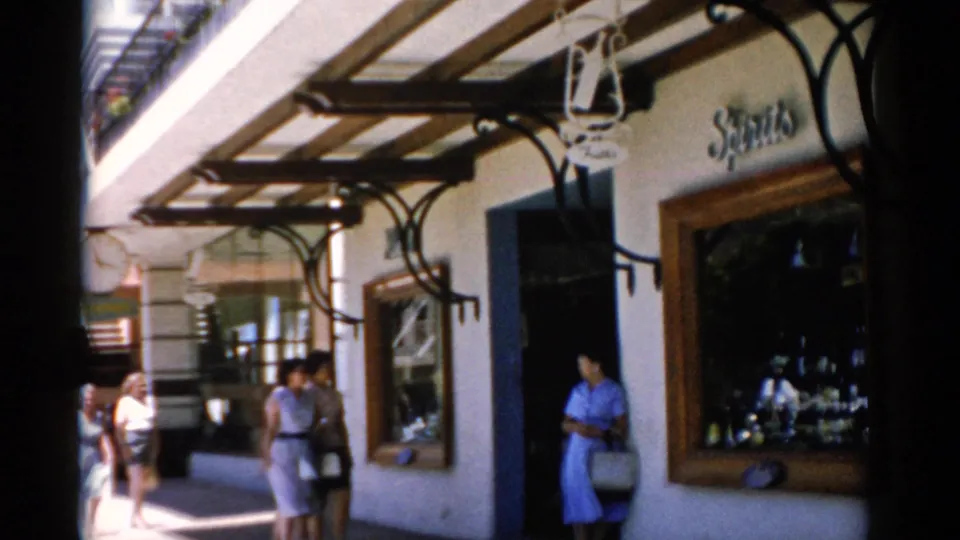 1975 Florida 8mm frame showing visitors outside a covered shopfront, with a woman in a blue dress near the entrance and signage above the window.