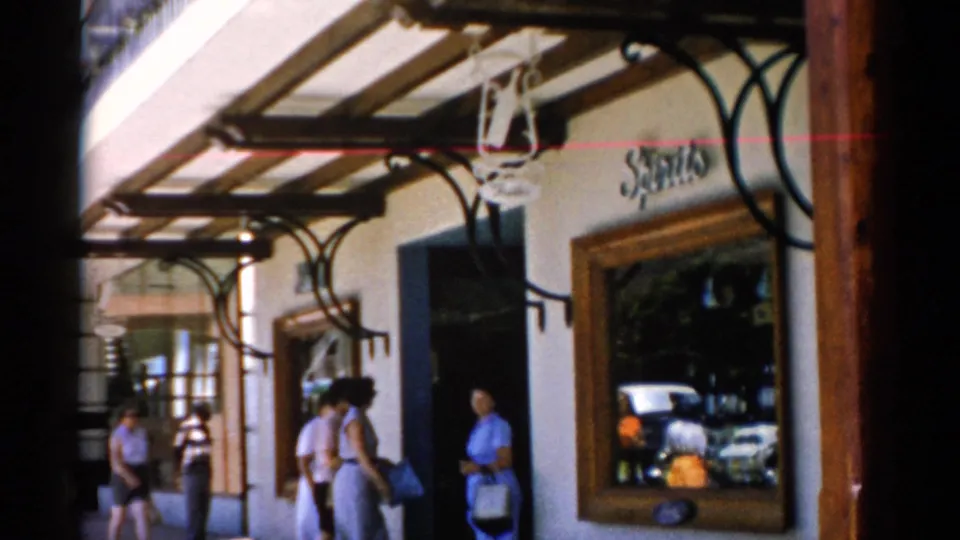 1975 Florida home-movie frame with pedestrians moving past a shop entrance, wooden awning supports, reflective display windows, and visible storefront signage.