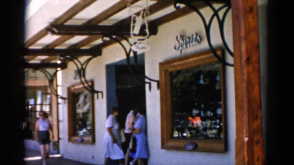 1975 Florida 8mm frame showing visitors near a shaded shop doorway, large display window, wooden awning post, and the Spirits sign above.