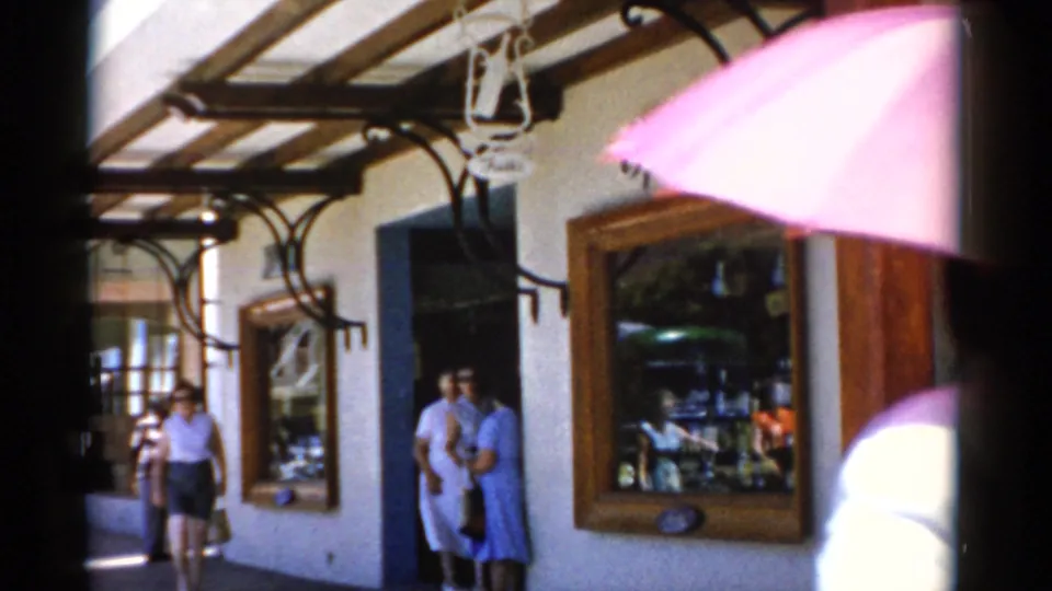 1975 Florida home-movie frame with two women outside a covered storefront, shop windows, wooden trim, and a pink umbrella partly blocking the upper right.