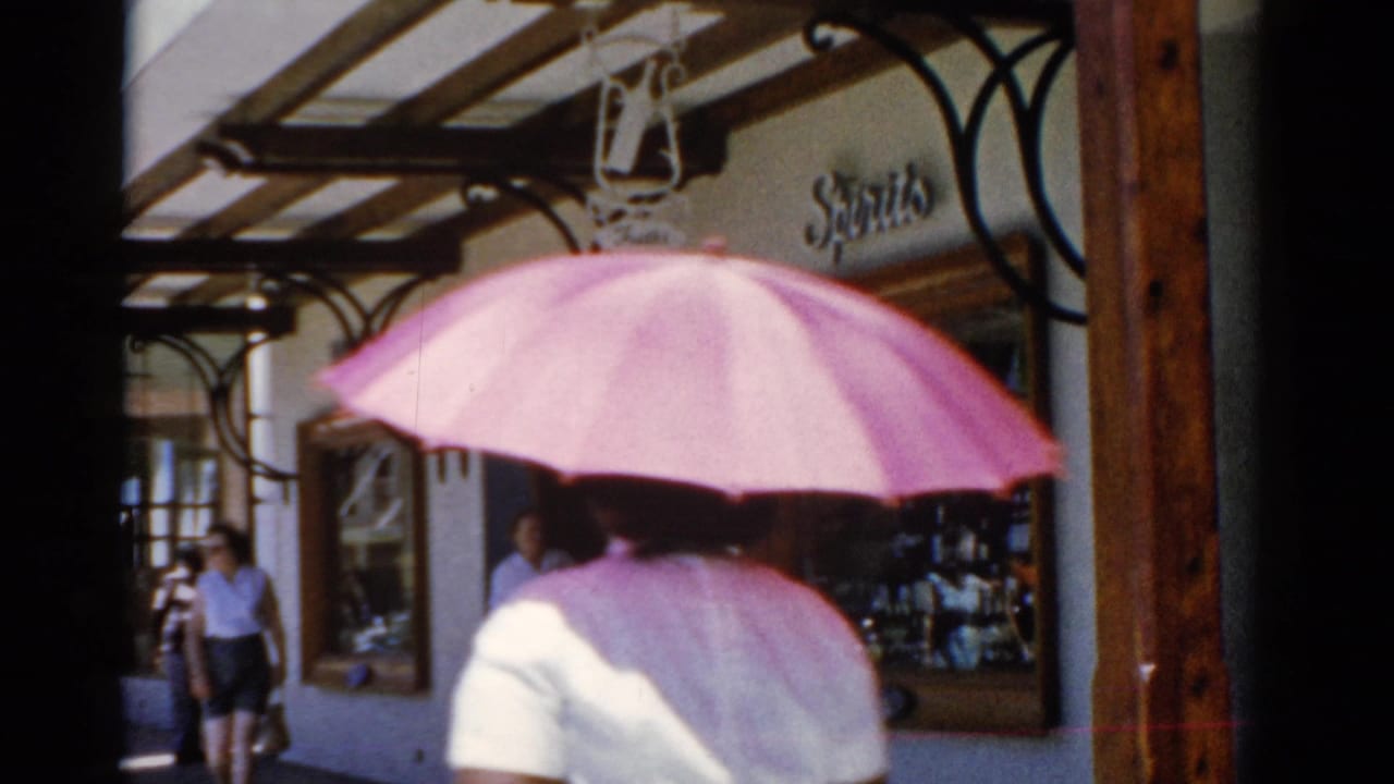 1975 8mm home-movie frame of a person with a pink umbrella walking past shops in a reported Florida travel scene.