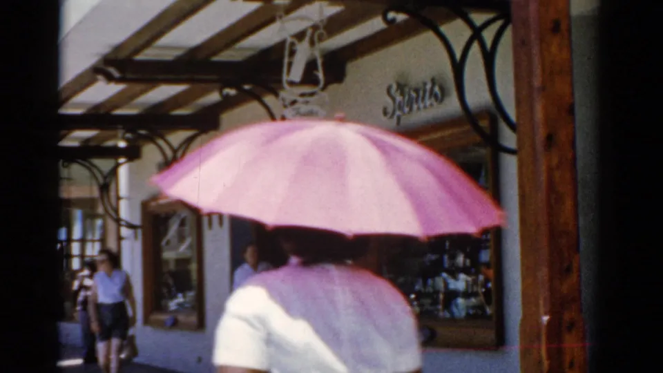 1975 8mm home-movie frame of a person with a pink umbrella walking past shops in a reported Florida travel scene.
