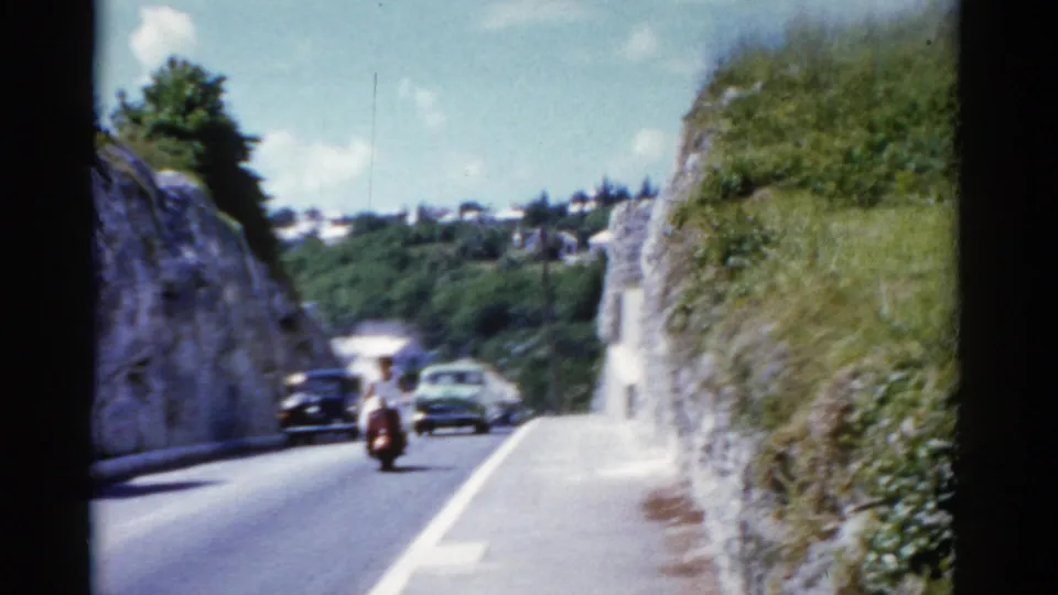 1970s 8mm travel footage frame of vehicles on a sunlit road bordered by rock walls and vegetation.