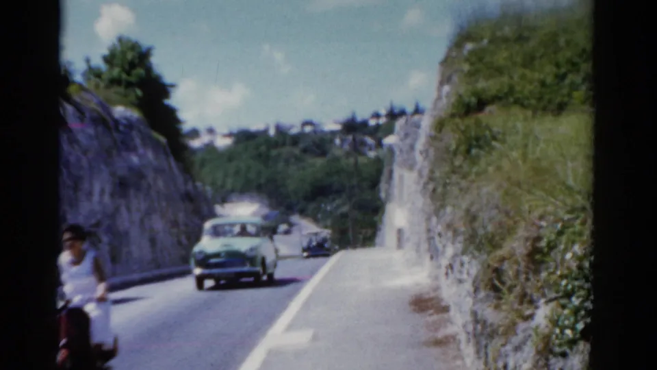 Reported Florida 1975 road scene with a classic car approaching through a rocky, green roadside landscape.
