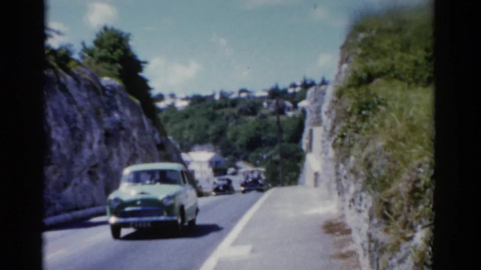 1975 Florida 8mm frame showing a light green period car on a narrow paved road bordered by rock faces and greenery.