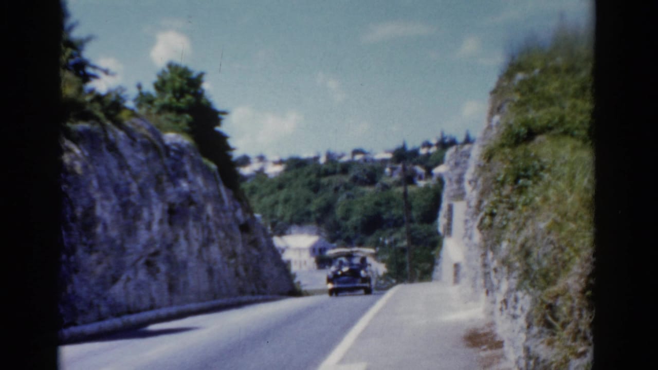1975 Florida 8mm frame with a dark period car moving through a narrow road cut, with hillside buildings beyond.