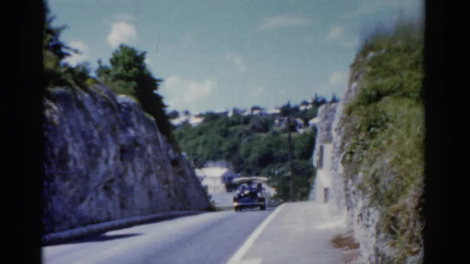 1975 Florida 8mm frame with a dark period car moving through a narrow road cut, with hillside buildings beyond.