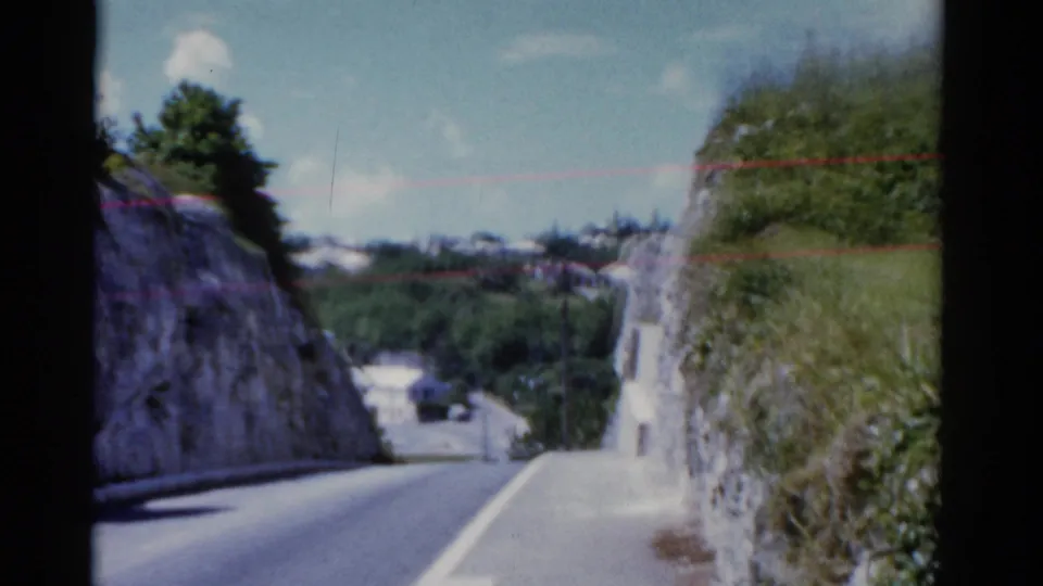 1975 Florida 8mm frame from a moving vehicle looking down a narrow road with period cars and rocky green slopes.