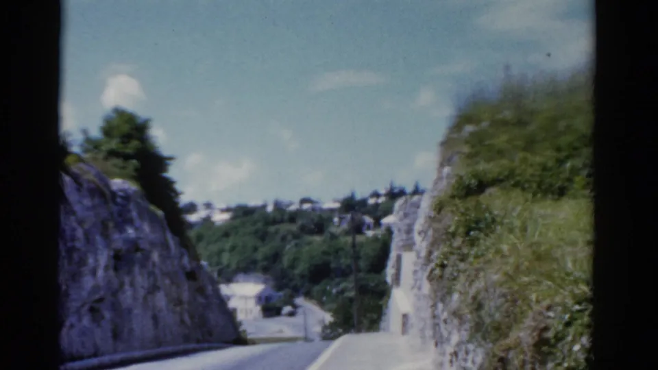 1975 Florida 8mm frame showing the road dropping between rocky green banks toward cars and white hillside structures.