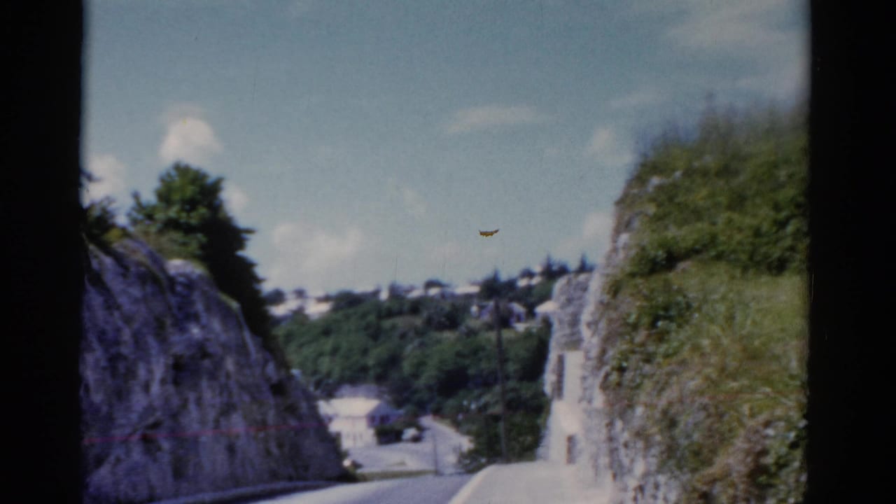 Forward-facing 8mm travel view along a road bordered by rock cuts and lush greenery, with distant structures visible beyond the slope.