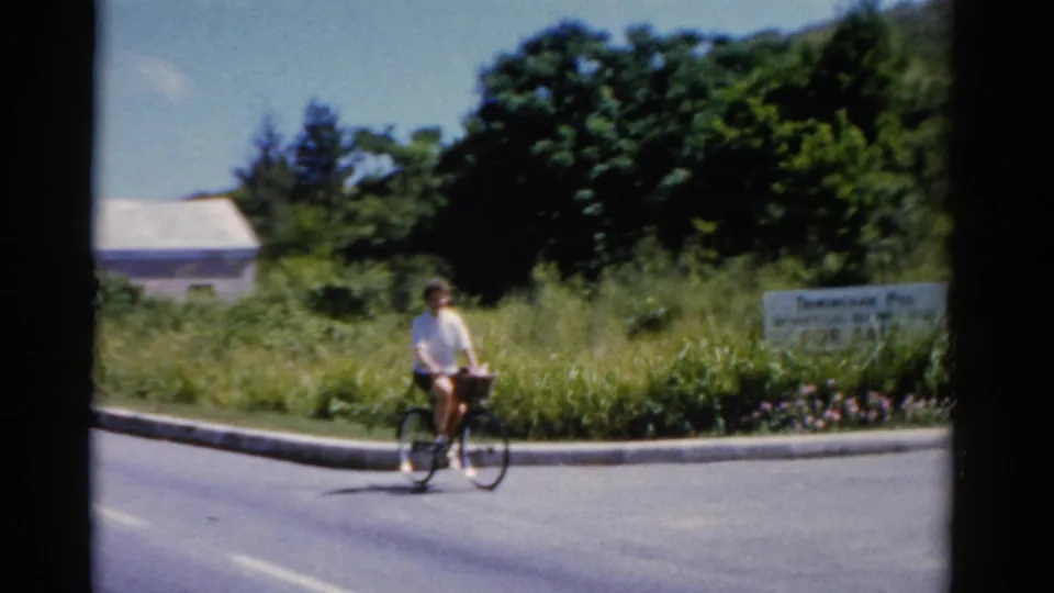 A person in a light shirt rides a bicycle along the roadside near a sign and dense vegetation in 1975 home-movie footage.