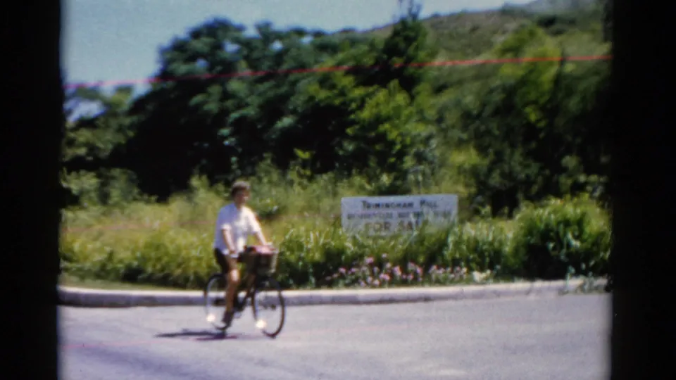 The cyclist rides past a roadside sale sign in a green roadside setting, with soft focus and visible 8mm film texture.