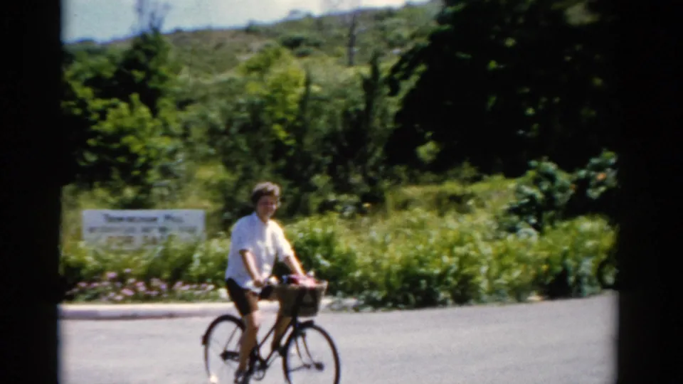 A woman in a white shirt and dark shorts rides a bicycle with a front basket beside lush Florida roadside vegetation.