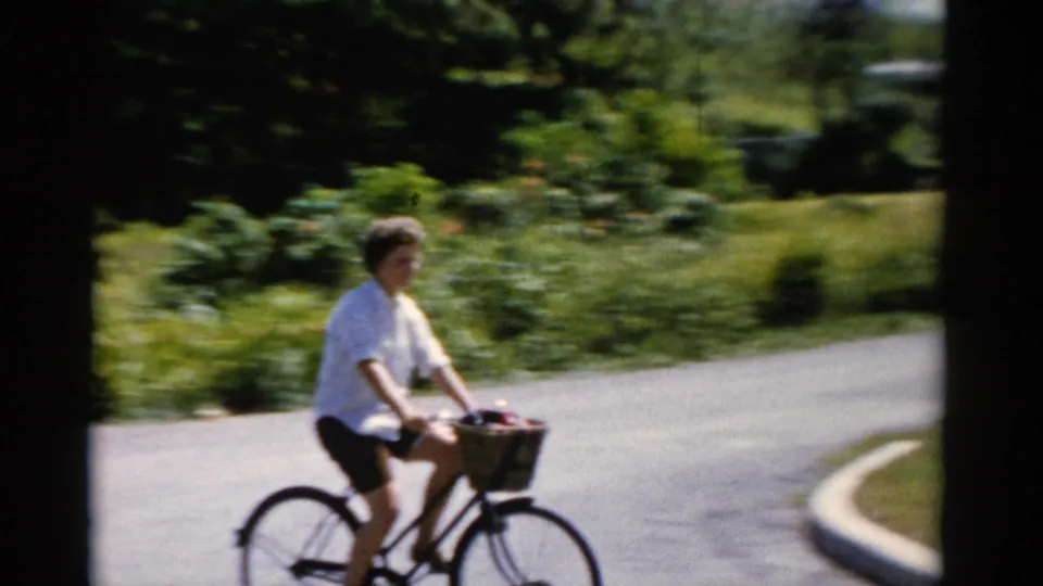 Closer 8mm view of the woman riding a bicycle with a front basket near a curved roadside bordered by grass and plants.