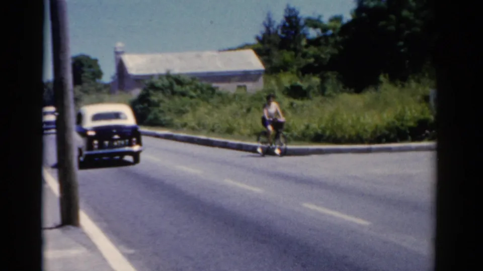 Wide 1975 Florida roadside view with a woman cycling near an intersection while an older car travels along the road.